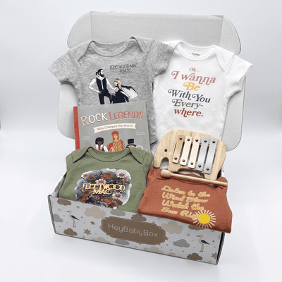 HeyBabyBox baby gift set featuring a curated collection of baby apparel and accessories, including a gray and white graphic bodysuit set, a green bodysuit, a rust-colored t-shirt, a wooden xylophone, and a 'Rock Legends' book, all arranged inside and in front of a gray cardboard box.