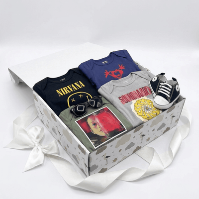 A baby gift set in a white patterned box with a white ribbon bow, containing a black Nirvana t-shirt, a blue baby bodysuit, a gray Soundgarden t-shirt, a green sweatshirt with a red graphic, a pair of black sunglasses, and a small black-and-white sneaker.