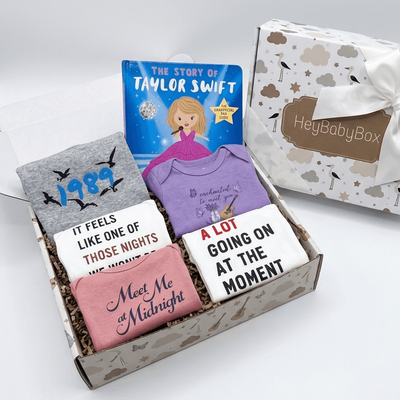A baby gift box set by HeyBabyBox containing a gray t-shirt with '1989', a purple t-shirt, a pink t-shirt with 'Meet Me Midnight', and two white t-shirts with humorous quotes, alongside a 'The Story of Taylor Swift' book and a branded gift box.
