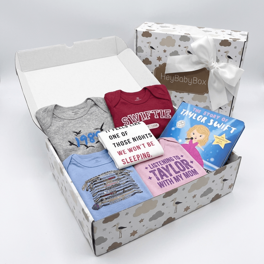 A baby gift box set from HeyBabyBox featuring a curated collection of baby apparel and a children's book. The open box contains a heather gray long-sleeve bodysuit with a blue bird graphic, a burgundy short-sleeve bodysuit with 'SWIFLE' text, a light blue striped shirt, a pink t-shirt, a white graphic sweatshirt, and a blue 'The Story of Taylor Swift' book.