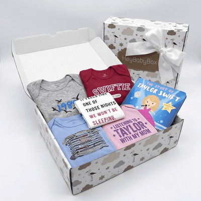 A baby gift box set from HeyBabyBox featuring a curated collection of baby apparel and a children's book. The open box contains a heather gray long-sleeve bodysuit with a blue bird graphic, a burgundy short-sleeve bodysuit with 'SWIFLE' text, a light blue striped shirt, a pink t-shirt, a white graphic sweatshirt, and a blue 'The Story of Taylor Swift' book.