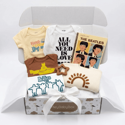 A baby gift box set featuring Beatles-themed apparel and accessories, including a pale yellow short-sleeve bodysuit, a white long-sleeve bodysuit with 'ALL YOU NEED IS LOVE' text, a brown short-sleeve bodysuit, a cream knit sun hat, a white star-shaped cookie, and a Beatles album book, arranged in an open box with tissue paper.