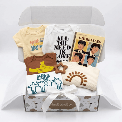 A baby gift box set featuring Beatles-themed apparel and accessories, including a pale yellow short-sleeve bodysuit, a white long-sleeve bodysuit with 'ALL YOU NEED IS LOVE' text, a brown short-sleeve bodysuit, a cream knit sun hat, a white star-shaped cookie, and a Beatles album book, arranged in an open box with tissue paper.