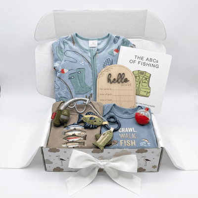 A baby fishing-themed gift box set containing a light blue patterned zip-up baby shirt, a blue 'Crawl Walk Fish' t-shirt, a wooden 'hello world' name card, a 'The ABCs of Fishing' booklet, fishing-themed accessories including a silver hook, fish-shaped pins, a small green lure, and a red bobber, all arranged in a white gift box with a white bow.