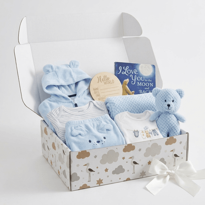 A baby gift box filled with soft blue baby clothing, a plush blue teddy bear, a small book, and a round wooden name sticker, set in a patterned box with a white ribbon bow on a light gray background.