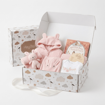 A baby gift set in a white cloud-patterned box, featuring a pink bunny hooded outfit, a plush bunny toy, a white baby t-shirt, and a book titled 'Bunny Roo'. A matching closed gift box labeled 'HeyBaby' sits behind the open box on a white background.