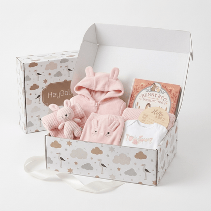 A baby gift set in a white cloud-patterned box, featuring a pink bunny hooded outfit, a plush bunny toy, a white baby t-shirt, and a book titled 'Bunny Roo'. A matching closed gift box labeled 'HeyBaby' sits behind the open box on a white background.