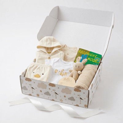 A baby gift box set containing a cream zip-up hooded romper, a white bodysuit with gold lettering, a knit duck toy, a beige knit blanket, a plush bunny, and a green book, all arranged inside a patterned gift box on a white background.