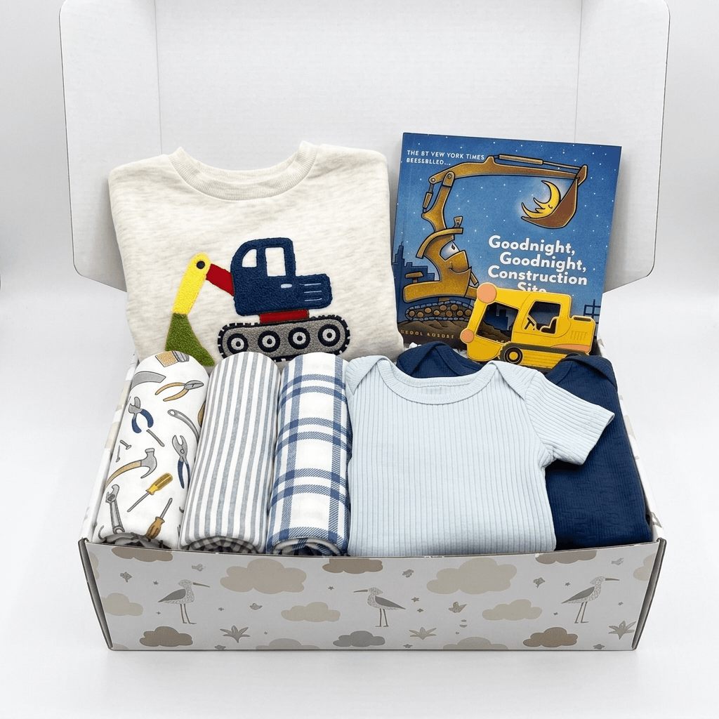 A baby gift box filled with baby clothing and a book. The set includes a cream t-shirt with a construction truck graphic, a blue and white striped blanket, a blue and white check blanket, a light blue ribbed bodysuit, a navy blue long-sleeve top, and a children's book titled 'Goodnight, Goodnight Construction Site'.