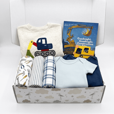 A baby gift box filled with baby clothing and a book. The set includes a cream t-shirt with a construction truck graphic, a blue and white striped blanket, a blue and white check blanket, a light blue ribbed bodysuit, a navy blue long-sleeve top, and a children's book titled 'Goodnight, Goodnight Construction Site'.