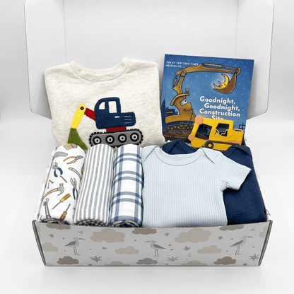 A baby gift box filled with baby clothing and a book. The set includes a cream t-shirt with a construction truck graphic, a blue and white striped blanket, a blue and white check blanket, a light blue ribbed bodysuit, a navy blue long-sleeve top, and a children's book titled 'Goodnight, Goodnight Construction Site'.