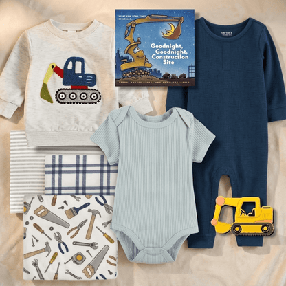 A baby construction-themed clothing set arranged on a beige fabric background, including a cream long-sleeve shirt with a blue and yellow excavator graphic, a navy one-piece, a light blue ribbed bodysuit, striped and plaid fabric pieces, a tool-print square, and a book titled 'Goodnight, Goodnight, Construction Site.'.