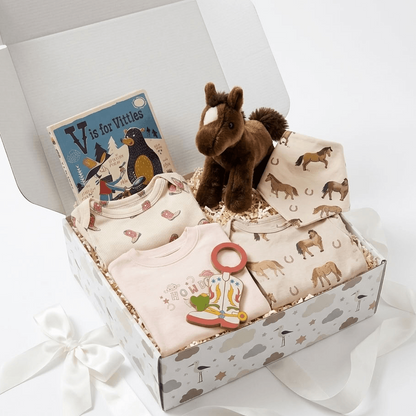 A baby gift box set with horse-themed items, including a plush brown horse toy, a patterned muslin blanket, a light pink baby t-shirt, a matching patterned bib, a small book, and a boot-shaped charm, all arranged inside an open patterned gift box with a white ribbon bow.