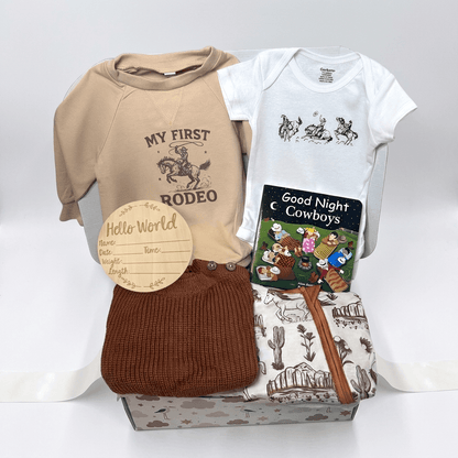 A baby gift set arranged on a white background, including a beige short-sleeve shirt with 'MY FIRST RODEO' text and graphic, a white short-sleeve bodysuit with cowboy illustrations, a brown knit hat, a folded cream blanket with western print, a small 'Hello World' wooden name card, and a 'Good Night Cowboys' book.