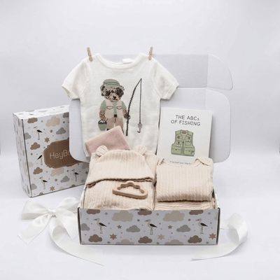 A baby gift set arranged in a white box with a matching cloud-patterned box and ribbon. The set includes a cream short-sleeve baby tee with a dog fishing graphic, a folded pale pink blanket, two folded cream ribbed baby garments in the front box, a small wooden toy, and a card reading 'THE ABC.
