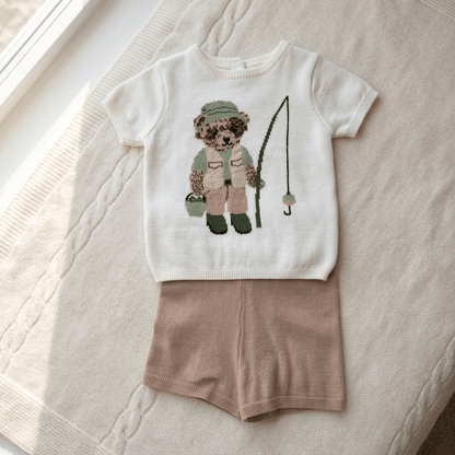 A baby boy's knit outfit consisting of a cream short-sleeve shirt with a teddy bear fishing graphic and matching dusty pink knit shorts, laid flat on a textured cream blanket.