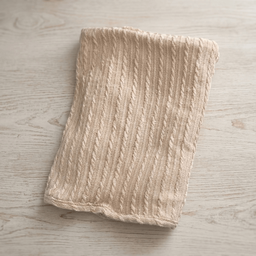A folded beige cable-knit fabric cloth, likely a tea towel or kitchen towel, displayed on a light wood grain surface.