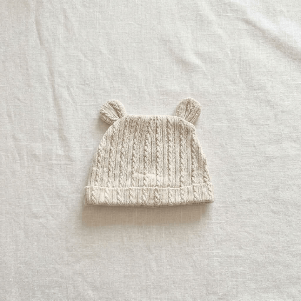 A cream-colored cable-knit baby hat with two small ear-like loops on top, laid flat on a textured off-white fabric background.
