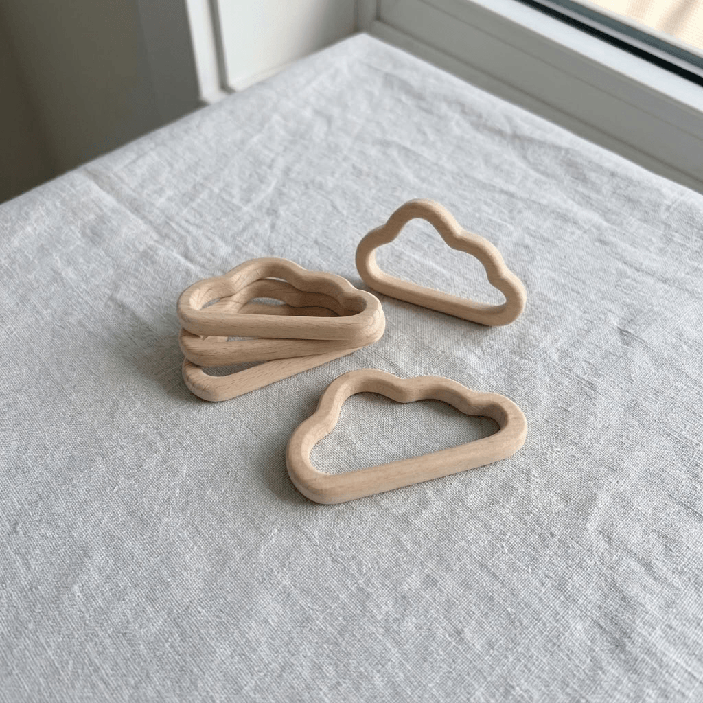 Three natural wood cloud-shaped baby teething rings arranged on a light gray linen cloth near a window.