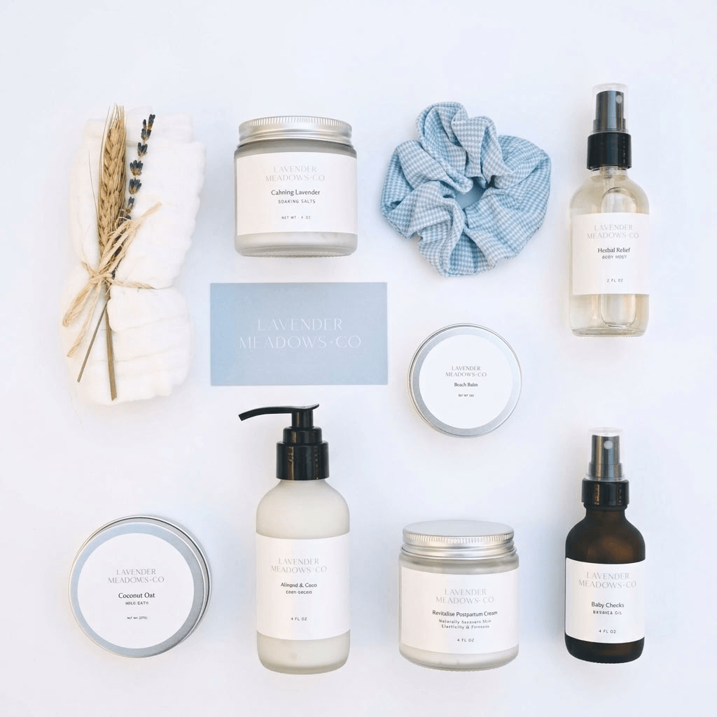A flat-lay arrangement of lavender-themed bath and body products from Lavender Meadows Co. on a white background, including a jar of body wash, a bottle of shower foam, a pump bottle of coconut oil, a jar of body butter, a small tin, a blue-and-white scrunchie, and dried lavender and wheat stems.