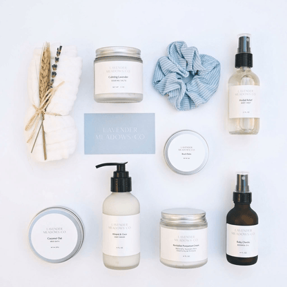 A flat-lay arrangement of lavender-themed bath and body products from Lavender Meadows Co. on a white background, including a jar of body wash, a bottle of shower foam, a pump bottle of coconut oil, a jar of body butter, a small tin, a blue-and-white scrunchie, and dried lavender and wheat stems.