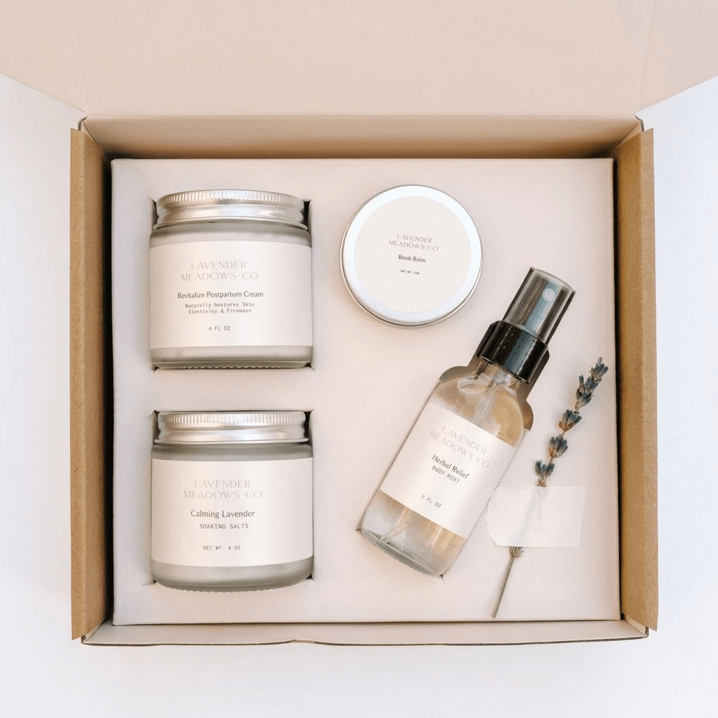 A curated wellness gift set from Lavender & Lemonade Co. arranged in a cardboard gift box, featuring two 'Calming Lemon' scented candles, a 'Body Butter' jar, a 'Handmade Hand Wash' spray bottle, and a sprig of lavender on a cream insert.