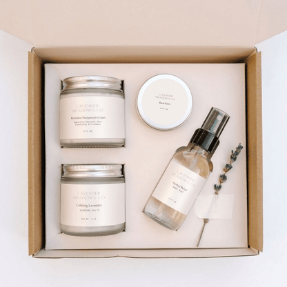 A curated wellness gift set from Lavender & Lemonade Co. arranged in a cardboard gift box, featuring two 'Calming Lemon' scented candles, a 'Body Butter' jar, a 'Handmade Hand Wash' spray bottle, and a sprig of lavender on a cream insert.