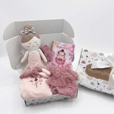 A baby ballet-themed gift set in an open white box, including a plush ballerina doll, pink tutu, ballet slippers, a small book, and a folded pink garment, alongside a branded HelloBabyBox gift box with a white bow.