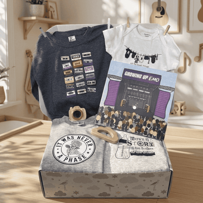A curated gift box set displayed on a wooden floor in a sunlit room. The set includes a navy graphic sweatshirt, a white graphic bodysuit, a folded light gray T-shirt with a circular 'It Was Never A Phase' patch, a printed card reading 'Growing Up EMO', and a gray patterned gift box.