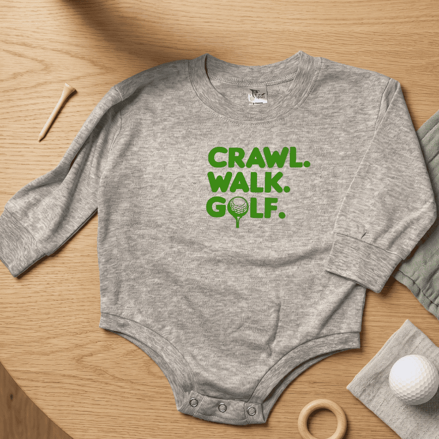 Gray baby onesie with 'CRAWL. WALK. GOLF.' text on a wooden surface with golf-themed items.