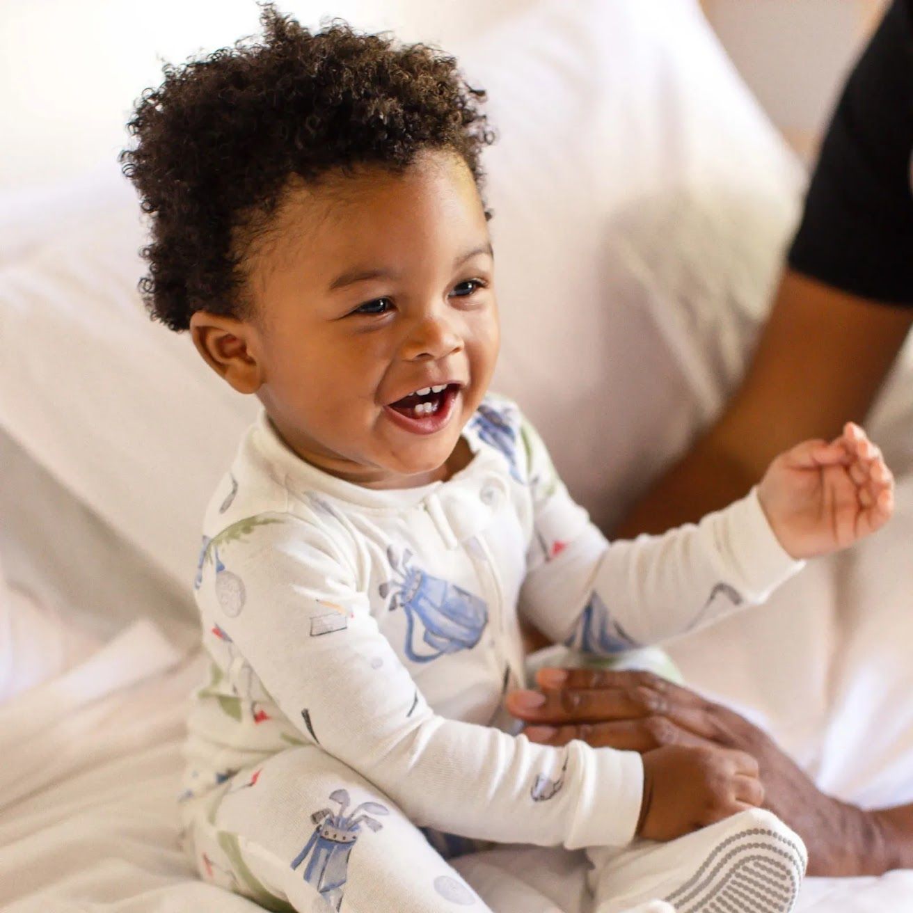 Baby in pajamas sitting on a bed with a person holding their hand