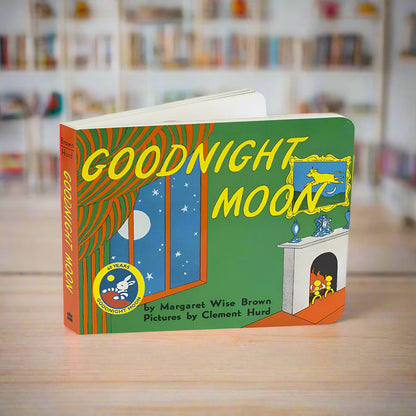 The children's board book 'Goodnight Moon' stands open on a light wooden table with a softly blurred library or bookstore background. The cover is green with large yellow title text, an orange striped curtain, blue moonlit window panels with white stars, and a white fireplace with yellow figures inside.