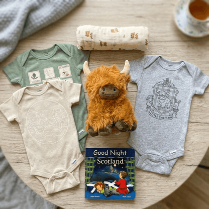 Baby clothing and plush toy on a wooden surface with a book titled 'Good Night Scotland'.