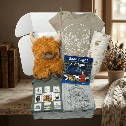 Collection of Scottish-themed baby items including t-shirts, a plush toy, and books on a wooden surface.