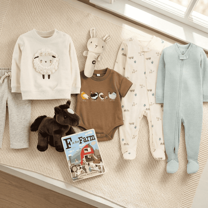 A styled flat-lay of baby clothing and accessories on a cream woven rug near a window. The set includes a cream sweatshirt with a sheep graphic, gray pants, a brown bodysuit with bird graphics, a cream footed one-piece with small prints, a light blue footed one-piece, a plush bunny toy, a plush dark brown horse, and a children's book titled 'Farm to My Farm'.