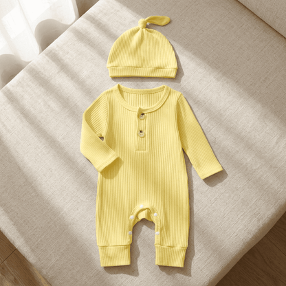 A baby's long-sleeve ribbed romper in pale yellow with a matching knotted beanie hat, laid flat on a textured beige fabric surface.