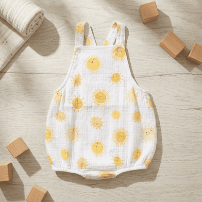 A white muslin baby romper with a yellow sun print, laid flat on a light wood surface with wooden blocks and a rolled blanket nearby.