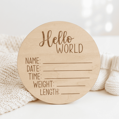 A round natural wood baby milestone plaque engraved with 'Hello WORLD' and lines for writing name, date, time, weight, and length. It is styled on a white surface next to a cream knit blanket and white knit socks.