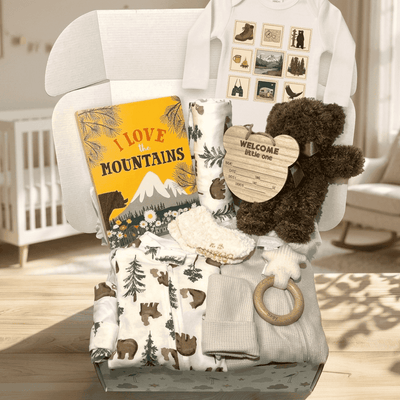 A curated baby gift set arranged in a white baby carrier on a wooden table in a softly lit nursery. The set includes a white long-sleeve bodysuit with a nature-themed print, a yellow 'I Love the Mountains' book, a dark brown plush teddy bear, a heart-shaped wooden tag, a white knit blanket, a wooden ring, and a folded gray blanket.