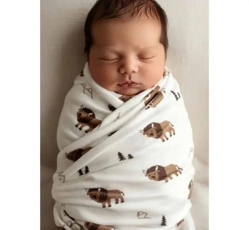 Hey Baby Box white swaddle blanket with a repeating pattern of cartoon bison and pine trees, wrapping a sleeping newborn baby.