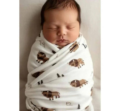 Hey Baby Box white swaddle blanket with a repeating pattern of cartoon bison and pine trees, wrapping a sleeping newborn baby.