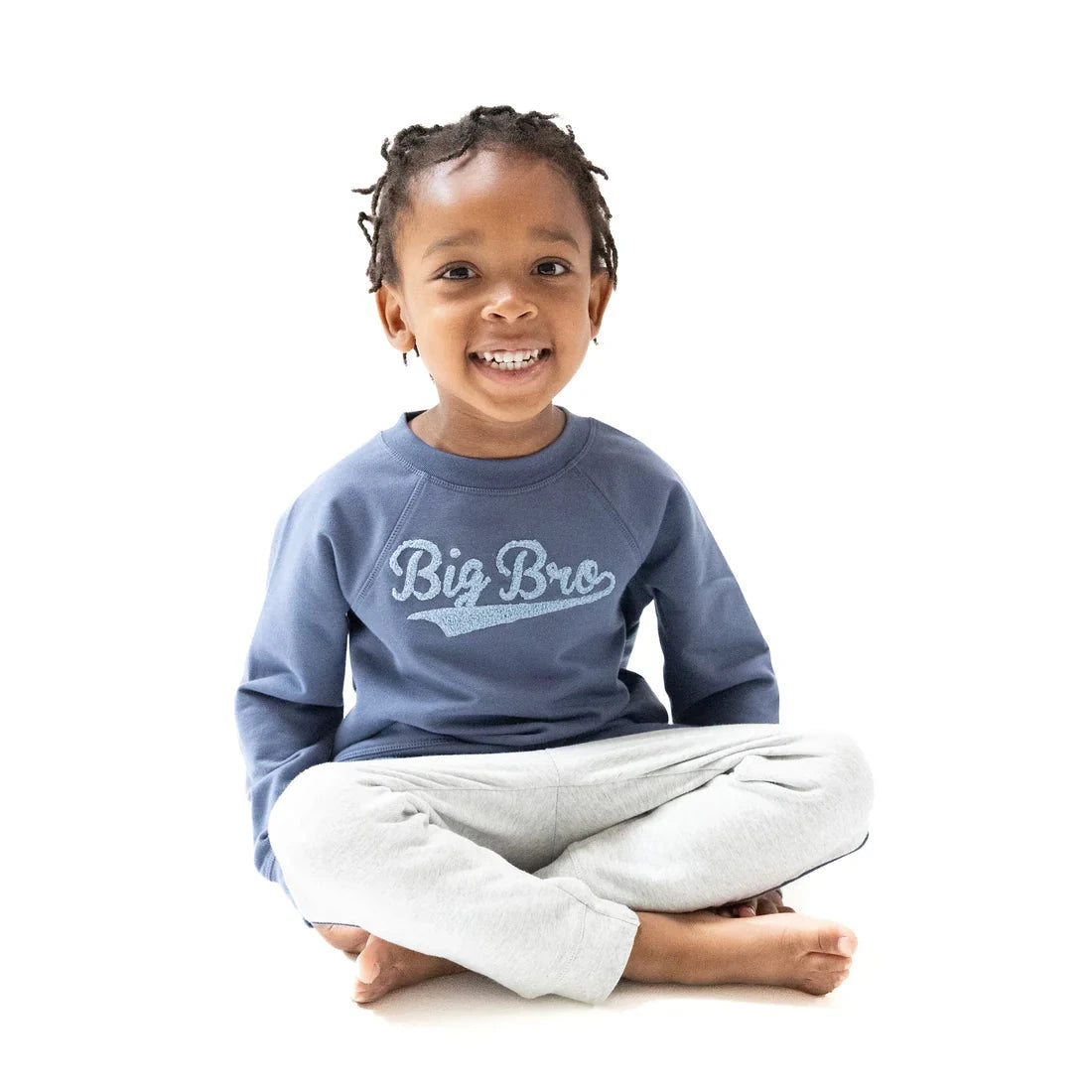 Child wearing a blue 'Big Bro' sweatshirt sitting cross-legged on a white background