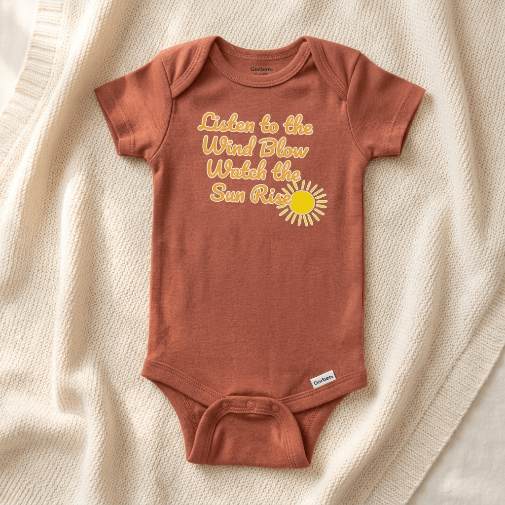 A terracotta short-sleeve baby bodysuit laid flat on a cream knit blanket. The bodysuit features a yellow graphic text that reads 'Listen to the Wind Blow Watch the Sun Rise' alongside a yellow sun illustration.