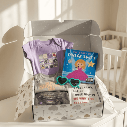A baby gift box filled with baby items, including a lavender t-shirt, a 'The Dream of Taylor Swift' book, a gray onesie, and a white comforter, displayed on a white dresser in a nursery setting.