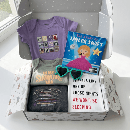 Box containing baby clothes and a book titled 'The Story of Taylor Swift' on a white surface.