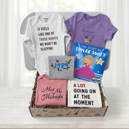 A curated baby gift box filled with personalized baby apparel and a children's book, arranged on a light wood surface in front of a soft white curtain background. The box contains a white long-sleeve bodysuit with black and red text, a purple short-sleeve bodysuit, a gray short-sleeve bodysuit with black bird graphics and '1984' text, a pink short-sleeve bodysuit with dark blue script text 