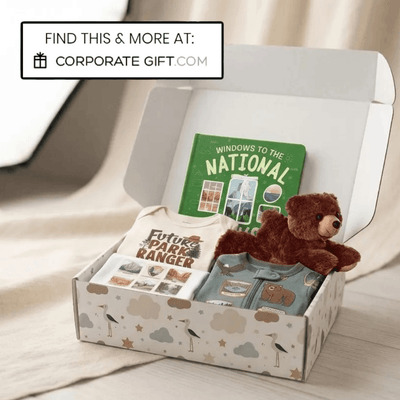 A curated gift box set displayed in an open white box with a cloud-patterned base. The set includes a green children's book titled 'Windows to the National', a white baby onesie printed with 'Future Park Ranger', folded blue and green clothing, and a small brown plush teddy bear.