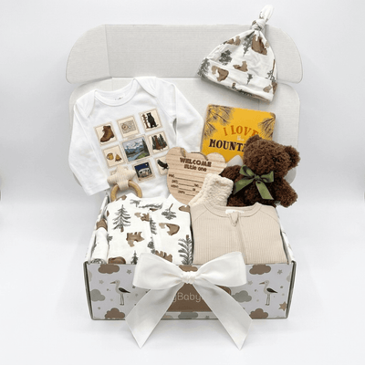 A baby gift box set containing a white long-sleeve bodysuit with a nature-themed photo print, a matching patterned hat, a brown teddy bear with a green bow, a cream ribbed top, patterned baby clothes, a wooden teething toy, and a yellow 'I Love Hounts' book, all arranged inside an open light gray box with a white ribbon bow.