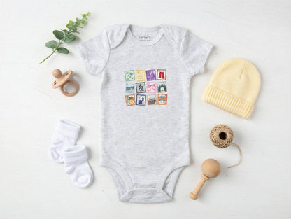 Baby onesie with colorful graphic, yellow hat, white socks, wooden rattle, and pacifier on a light background