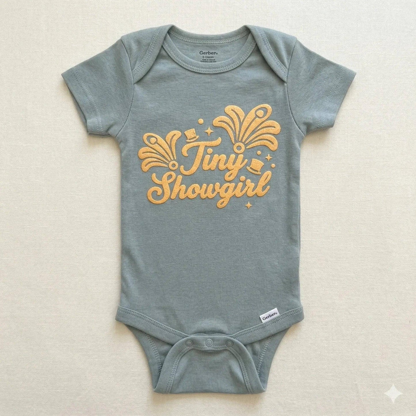 Gray baby onesie with 'Tiny Showgirl' text on a beige background